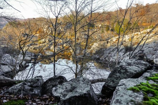 Looking down at the western rocky border of Black Pond in the distance with a boulder outcropping and leafless trees in the foreground.